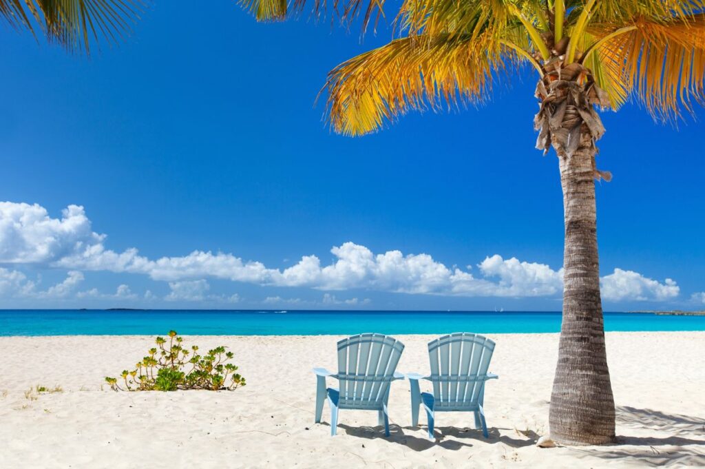 Two blue Adirondack chairs under a palm tree on a pristine sandy beach, overlooking a calm turquoise sea under a clear blue sky. This serene setting captures the exclusivity of Anguilla, inviting you to unwind in unparalleled tranquility.