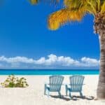 Two blue Adirondack chairs under a palm tree on a pristine sandy beach, overlooking a calm turquoise sea under a clear blue sky. This serene setting captures the exclusivity of Anguilla, inviting you to unwind in unparalleled tranquility.