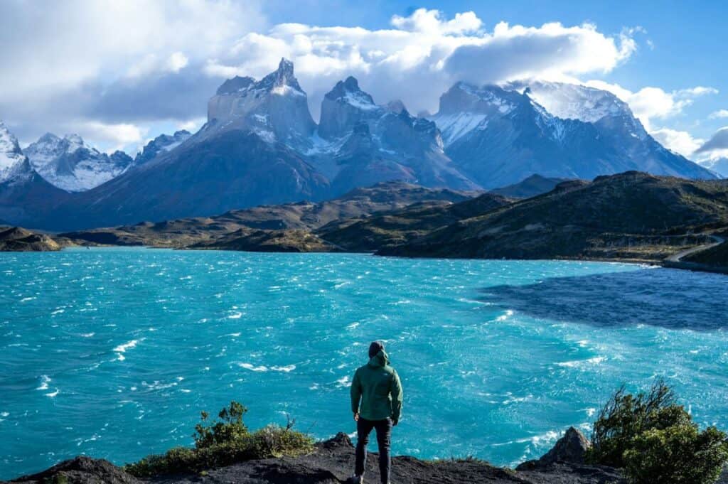 A person in a green jacket stands on a rock, overlooking a turquoise lake with a mountainous landscape in the background under a partially cloudy sky.