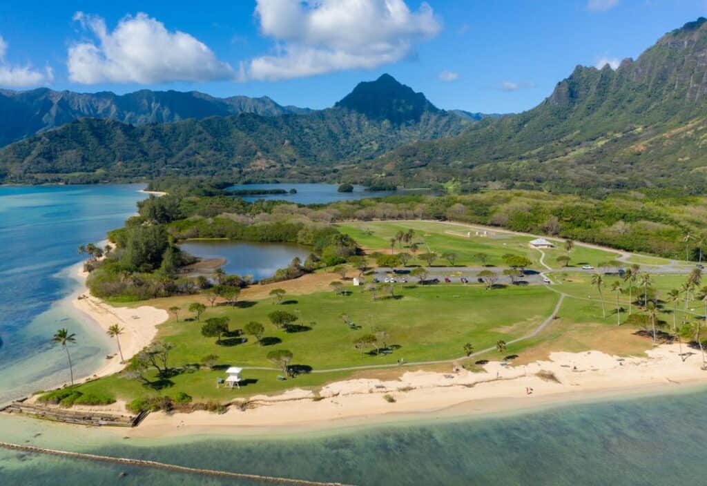 Aerial view of a coastal park with lush greenery, mountains in the background, sandy beaches, and a clear blue sky with scattered clouds.