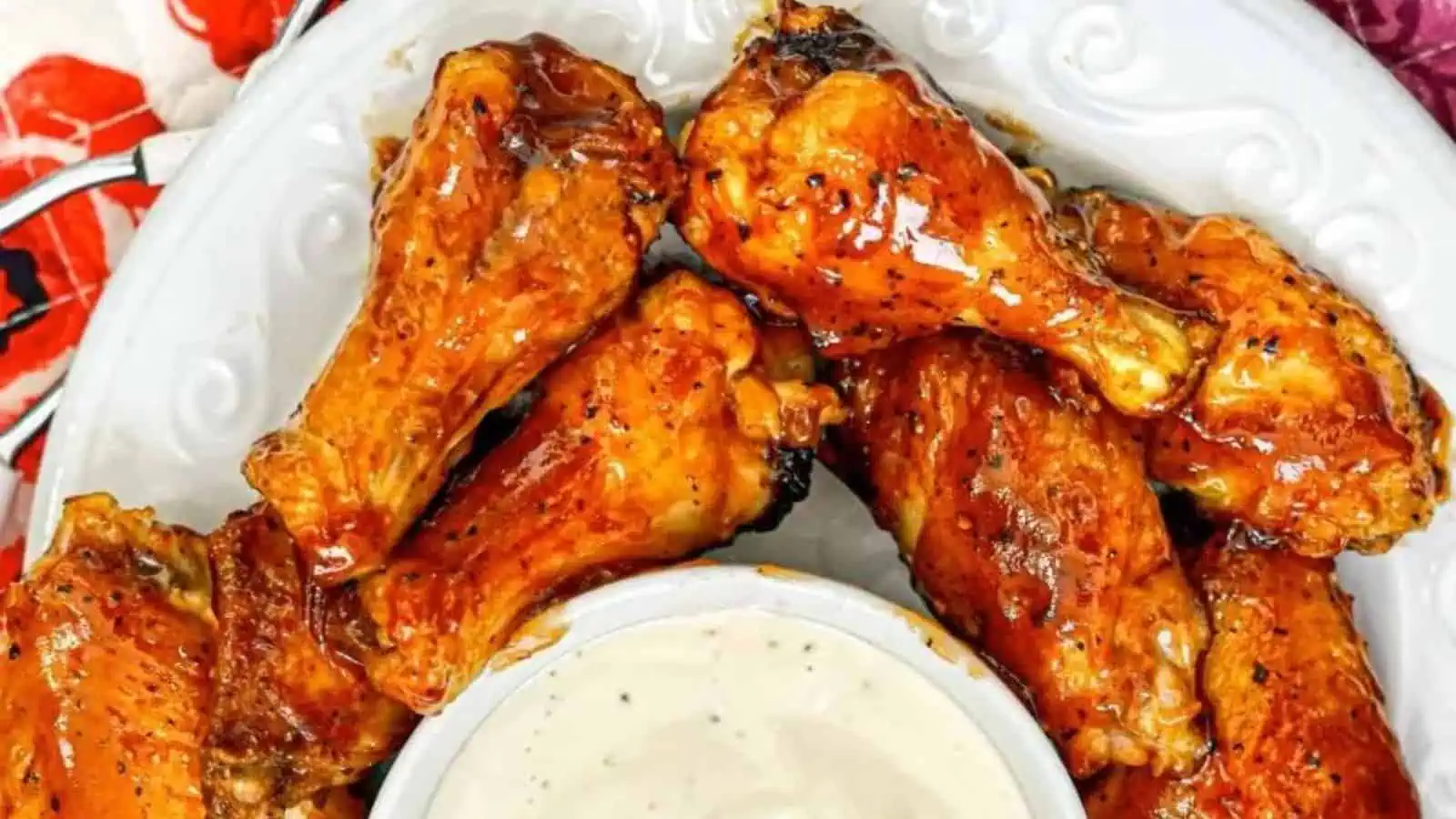 Chicken wings glistening with a rich, sticky barbecue sauce arranged on a white plate accompanied with a small bowl of creamy dipping sauce.