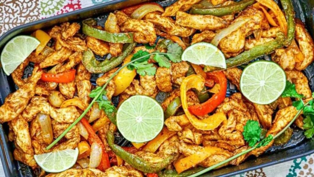 A tray of cooked chicken fajitas with sliced bell peppers, onions, lime wedges, and cilantro garnish.