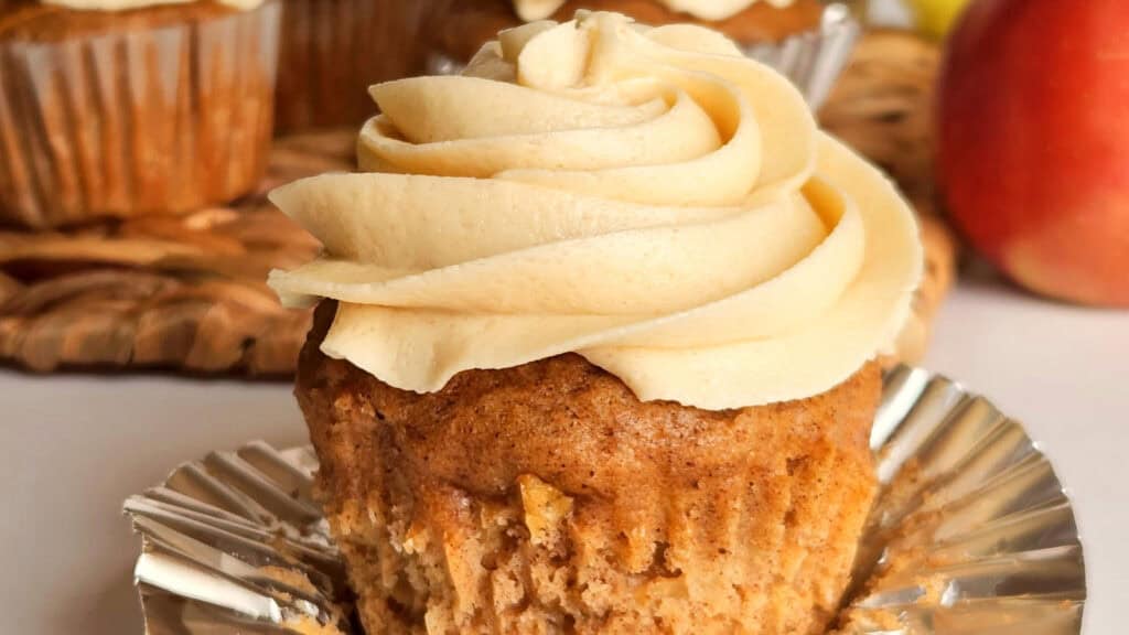 A close-up of a cupcake with a swirl of light brown frosting on top, partially unwrapped from its metallic foil liner. Another cupcake and a piece of wicker are visible in the background.