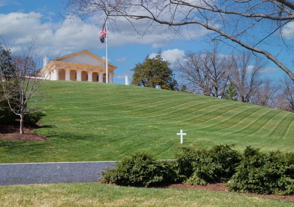 A white cross grave marker stands on a manicured lawn leading up to a large, columned building with an American flag, under a blue sky with clouds, reminiscent of the solemn beauty highlighted in Arlington National Cemetery facts.