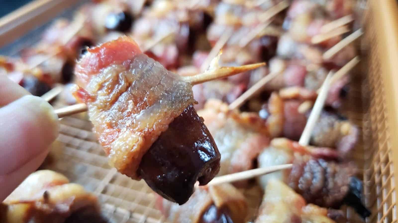 A close-up of a bacon-wrapped date held by a hand with a toothpick, with multiple bacon-wrapped dates on a tray in the background.