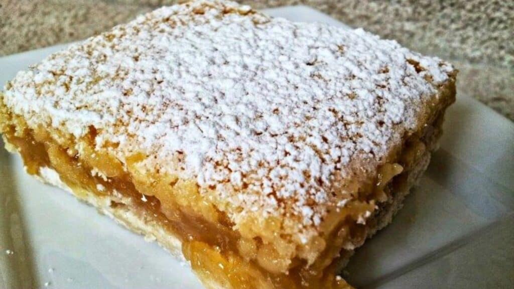 A close-up of a square slice of a lemon bar dessert, topped with a generous dusting of powdered sugar, served on a white plate.