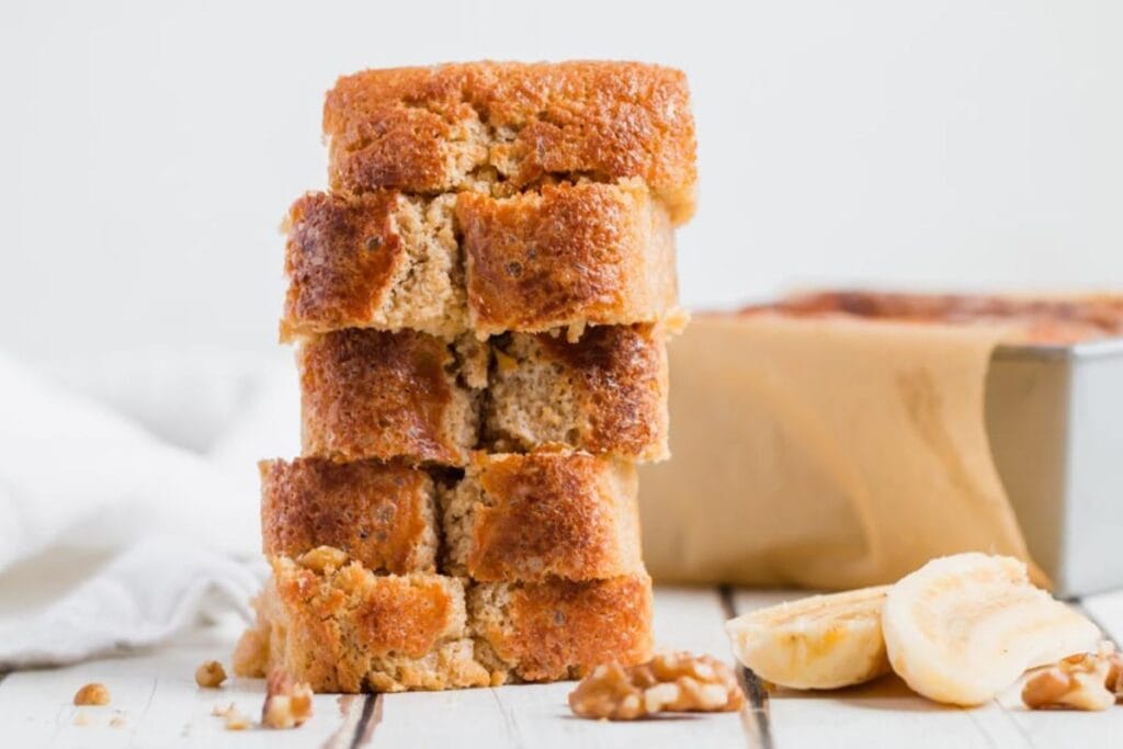 A tall stack of four slices of moist banana bread is positioned neatly next to sliced banana pieces and walnut halves on a white surface.