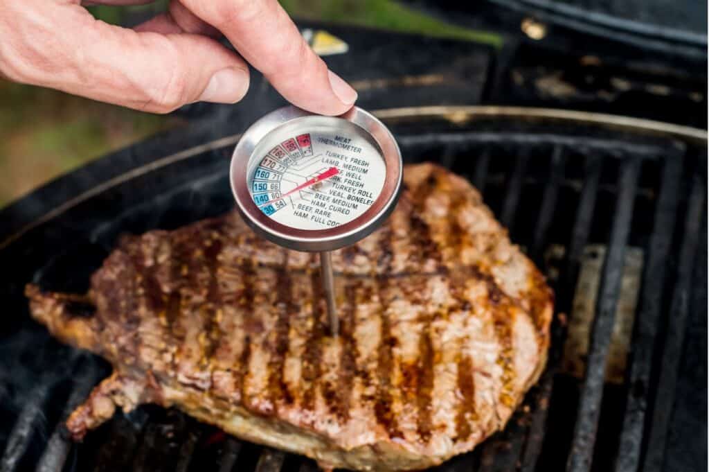 A hand holds a meat thermometer inserted into a steak on a grill, checking its temperature.