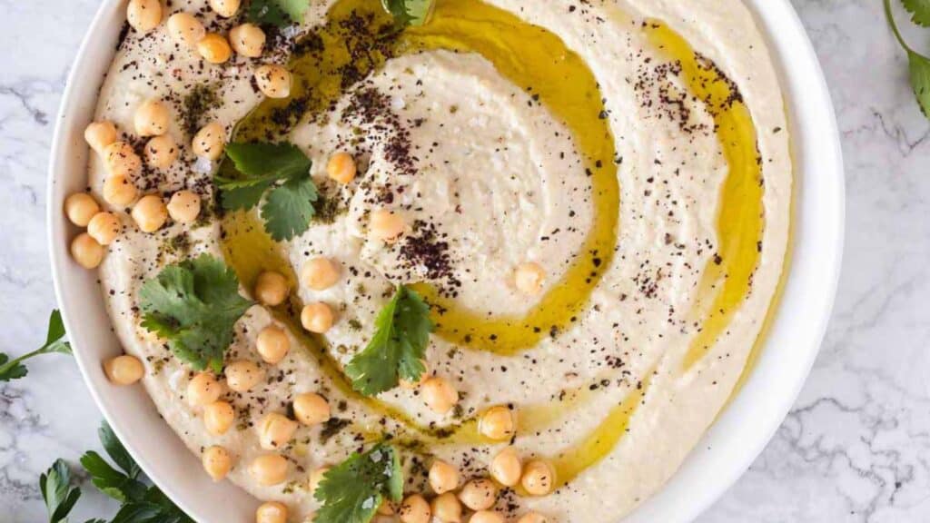 A bowl of hummus garnished with chickpeas, olive oil, parsley, and sprinkled spices on a marble surface.