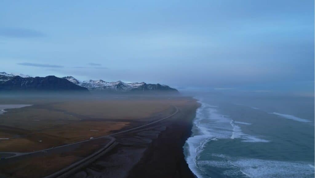 A coastal landscape with mist covering the shore, mountains in the distance, and water waves crashing against a rocky beach—a perfect snapshot for your 4-day Iceland itinerary. A winding road runs parallel to the shoreline.