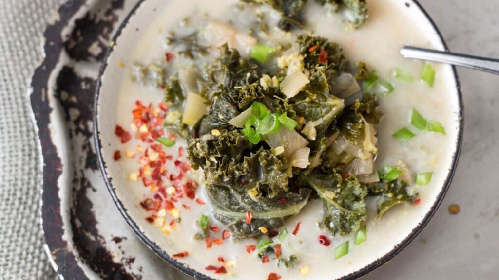 A bowl of soup topped with kale, chopped green onions, and red pepper flakes, with a spoon on the side.