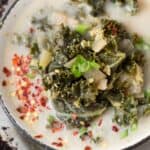 A bowl of soup topped with kale, chopped green onions, and red pepper flakes, with a spoon on the side.