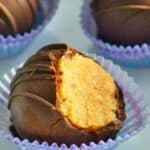 A close-up of a bitten chocolate-covered truffle in a purple wrapper, with two more unbitten truffles in the background, also in purple wrappers.