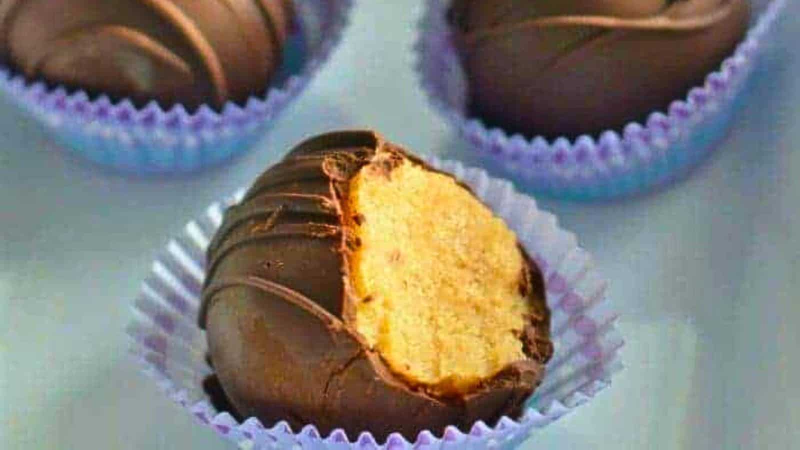 A close-up of a bitten chocolate-covered truffle in a purple wrapper, with two more unbitten truffles in the background, also in purple wrappers.