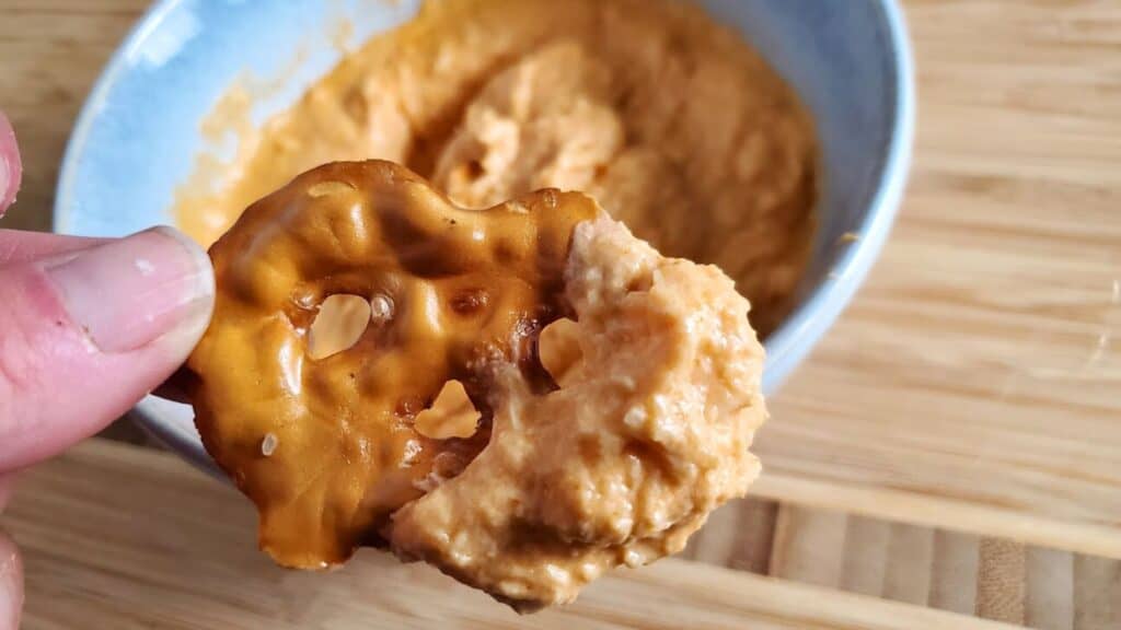 Close-up of a hand holding a pretzel chip dipped in a creamy, orange-colored cheese spread. A bowl of the buffalo chicken dip is in the background.