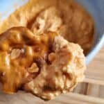 Close-up of a hand holding a pretzel chip dipped in a creamy, orange-colored cheese spread. A bowl of the buffalo chicken dip is in the background.