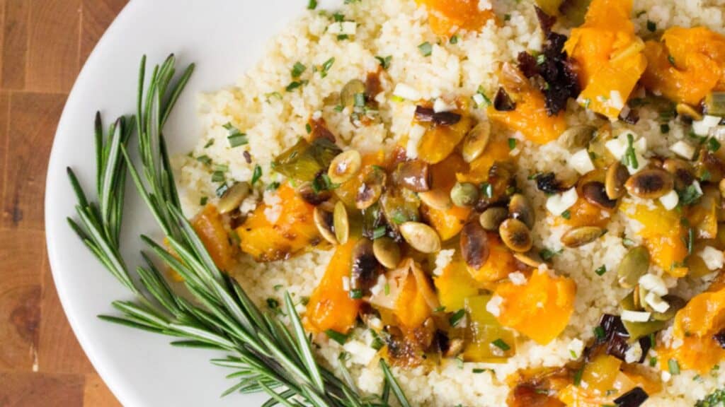 A white plate of couscous topped with roasted vegetables, pumpkin seeds, and garnished with a sprig of rosemary, placed on a wooden table.