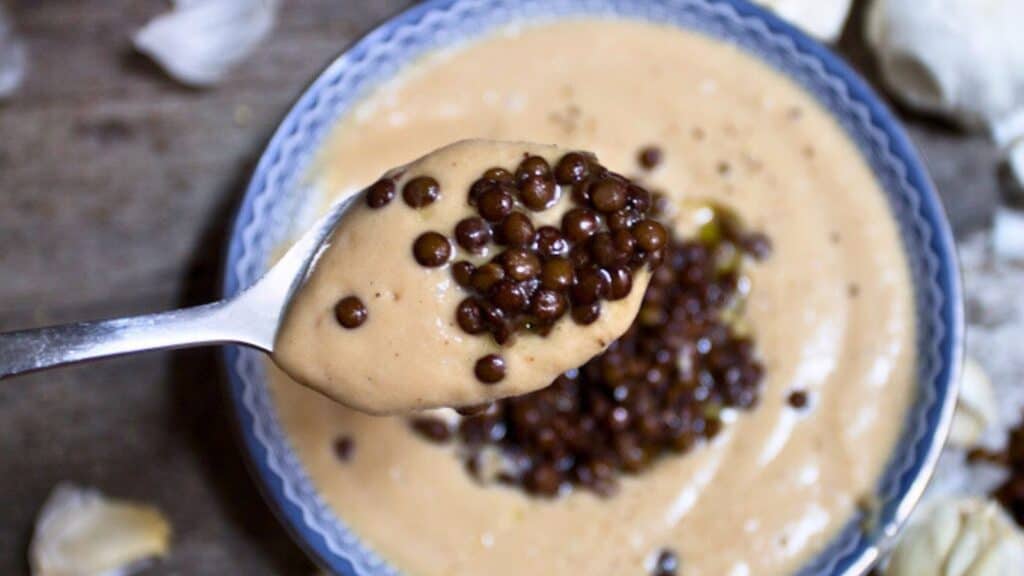 A bowl of creamy soup topped with black lentils, with a spoon lifting a portion of the soup and lentils.