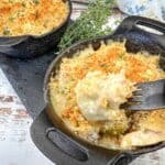 Two cast-iron skillets filled with creamy, baked casserole topped with golden breadcrumbs and garnished with fresh thyme sprigs, set on a wooden surface. A fork rests in one serving.