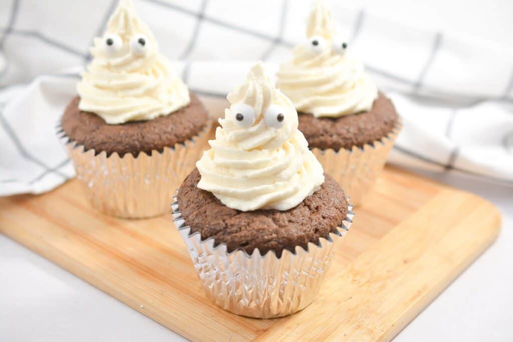 Three chocolate cupcakes with white frosting decorated to resemble ghosts, complete with googly eyes, placed on a wooden board against a checkered background.