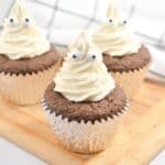 Three chocolate cupcakes with white frosting decorated to resemble ghosts, complete with googly eyes, placed on a wooden board against a checkered background.