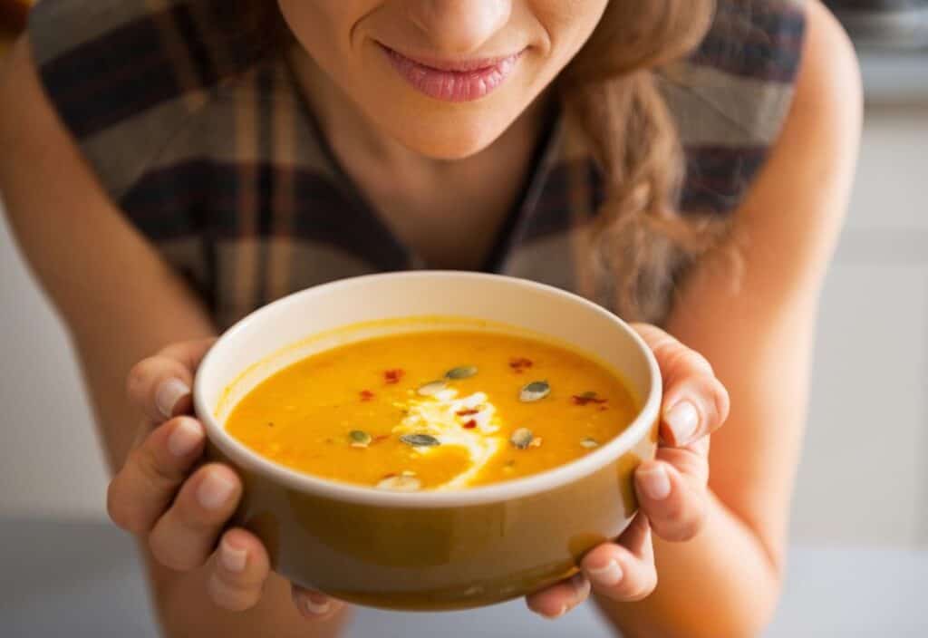 A person holds a bowl of creamy orange soup topped with seeds and a dollop of cream.