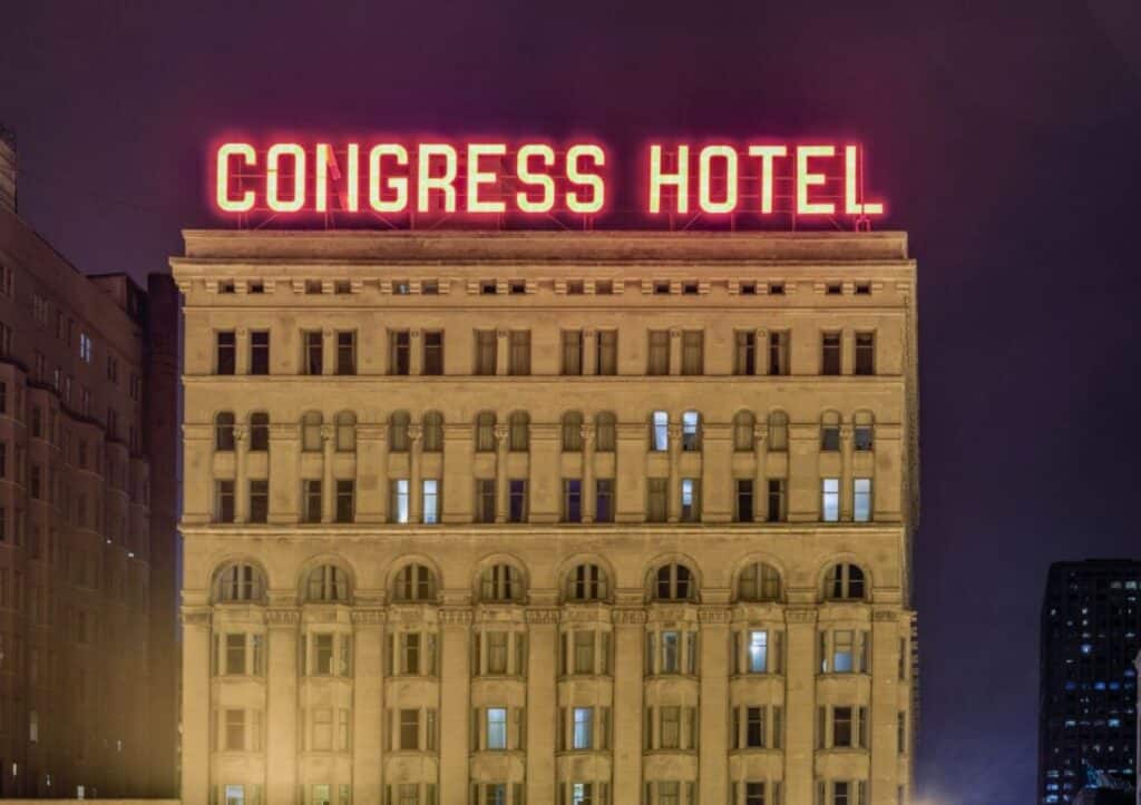 The exterior of Congress Hotel at night, with "CONGRESS HOTEL" illuminated in red neon lights on the roof of the building.