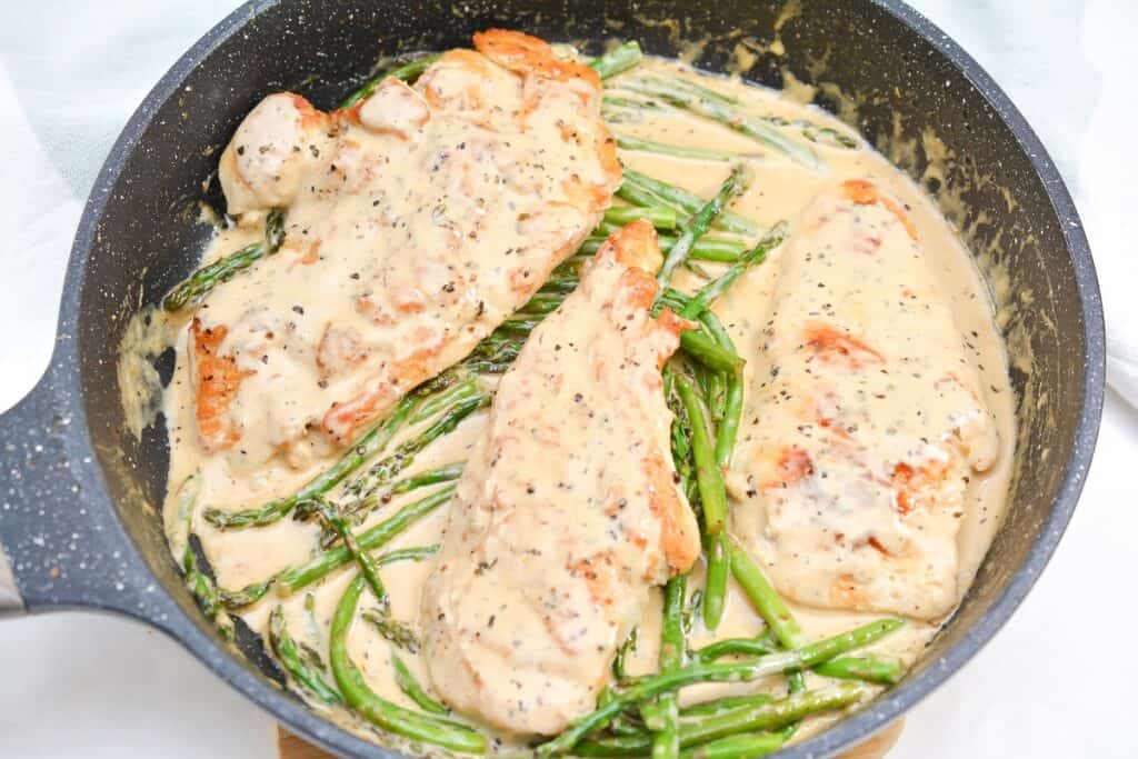 A skillet containing three chicken breasts and asparagus spears in a creamy sauce, seasoned with black pepper.