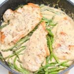 A skillet containing three chicken breasts and asparagus spears in a creamy sauce, seasoned with black pepper.