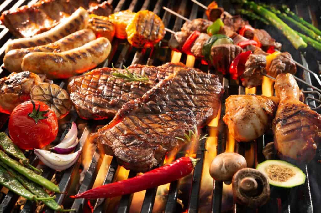 A variety of meats and vegetables are being grilled on a barbecue, including sausages, steak, chicken drumsticks, bell peppers, tomatoes, mushrooms, zucchini, garlic, and asparagus. For grilling tips for beginners: ensure even cooking by maintaining consistent heat and not overcrowding the grill.