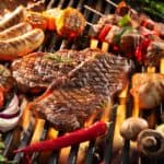 A variety of meats and vegetables are being grilled on a barbecue, including sausages, steak, chicken drumsticks, bell peppers, tomatoes, mushrooms, zucchini, garlic, and asparagus. For grilling tips for beginners: ensure even cooking by maintaining consistent heat and not overcrowding the grill.