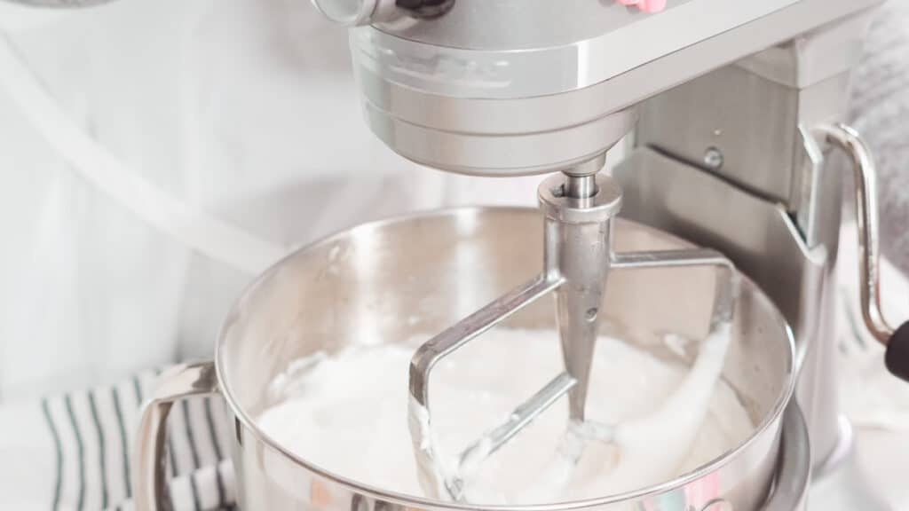 A stand mixer with a paddle attachment is mixing a white, fluffy substance in a stainless steel bowl.