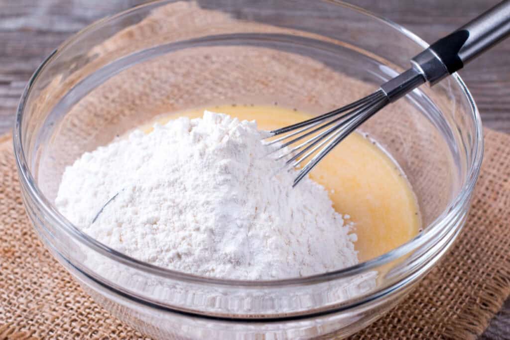 A glass bowl containing flour and a whisk on top of a mixture of wet ingredients. The bowl is placed on a burlap cloth on a wooden surface.