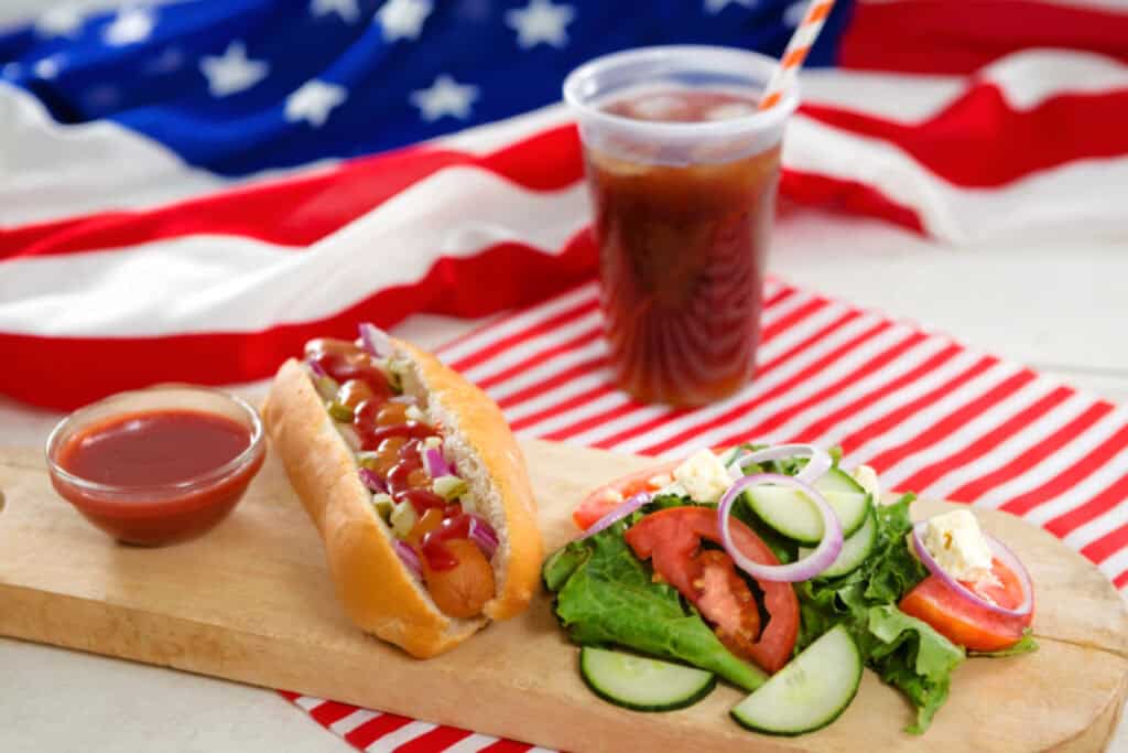 A hot dog with relish and mustard on a wooden board next to a small bowl of ketchup, a side salad, and a cup of iced tea. An American flag is in the background.