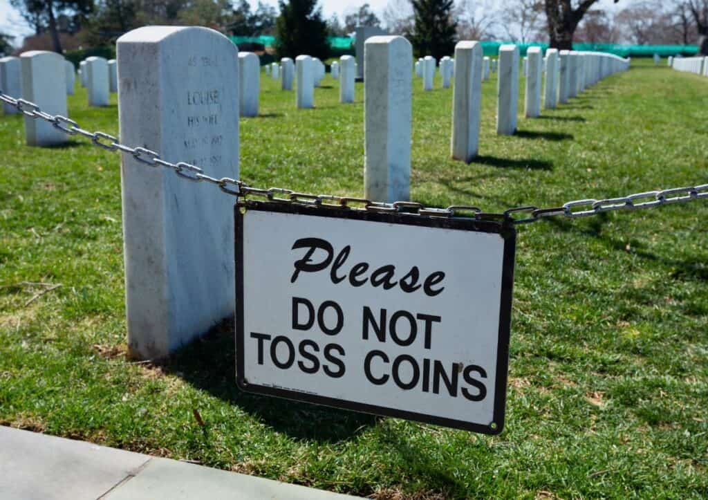 A sign in a cemetery reads "Please Do Not Toss Coins," with gravestones arranged in rows in the background, reminiscent of Arlington National Cemetery's solemn arrangement.