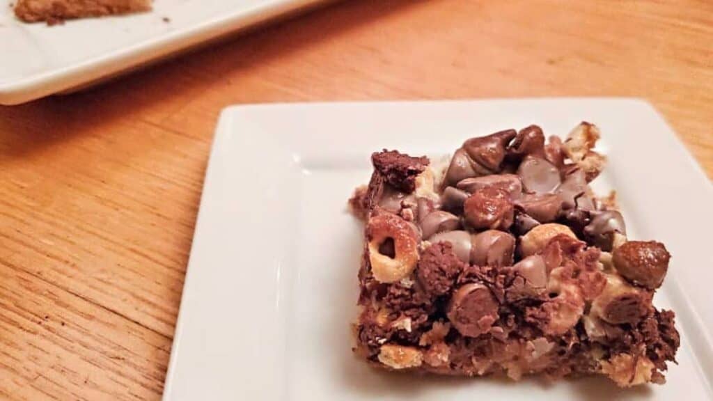 A dessert bar topped with chocolate chips and cereal pieces is placed on a white square plate on a wooden table. Another plate with a dessert bar is partially visible in the background.