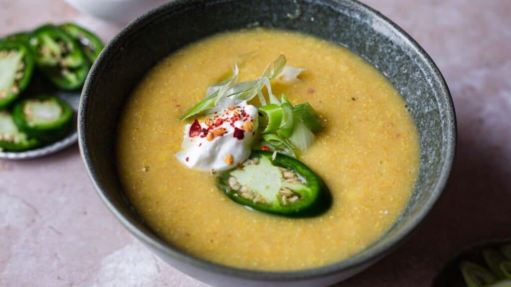 A bowl of creamy soup garnished with sliced jalapeño, a dollop of sour cream, chopped green onions, and red pepper flakes, with a small plate of sliced jalapeños beside it.