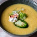 A bowl of creamy soup garnished with sliced jalapeño, a dollop of sour cream, chopped green onions, and red pepper flakes, with a small plate of sliced jalapeños beside it.