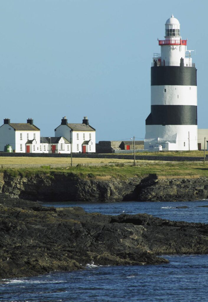 A black and white striped lighthouse stands on a grassy area near three white cottages with red doors by a rocky shoreline, the perfect picturesque spot to start your 4-day Ireland itinerary. The sky is clear and blue.