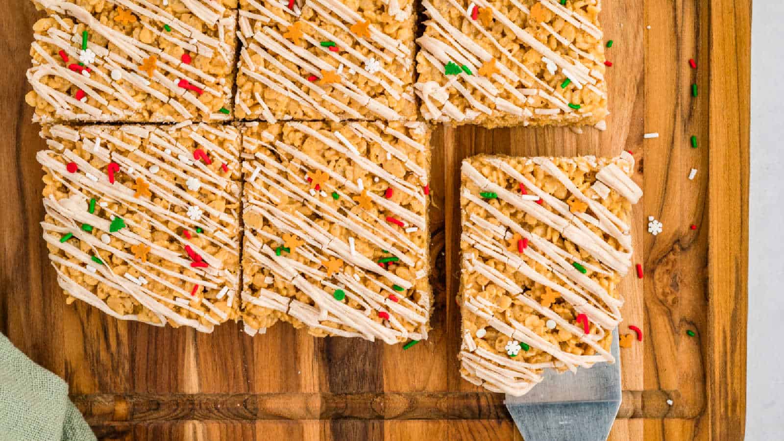 Rice Krispies treats with white frosting drizzle and festive sprinkles, cut into squares on a wooden cutting board, with one square being lifted by a spatula.