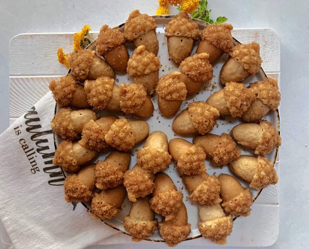 A plate filled with acorn-shaped cookies, with brown tops resembling acorn caps and a lighter base. The plate is placed on a white surface, partially covered by a napkin.