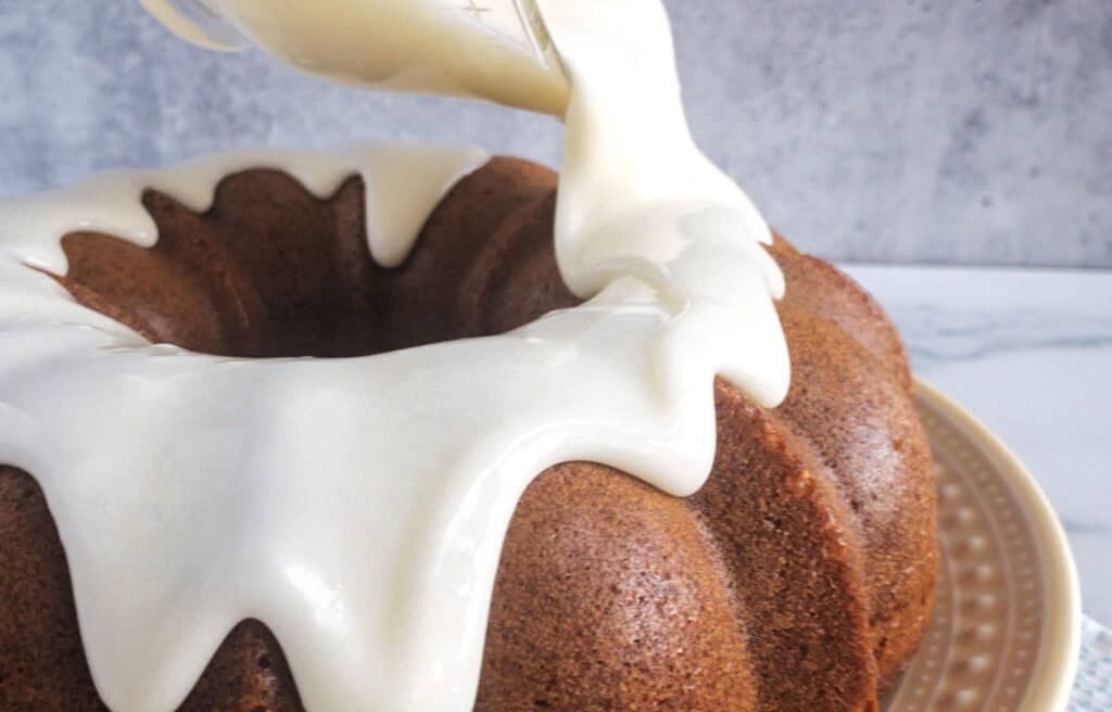 Close-up of a Bundt cake being drizzled with thick white icing, set on a beige plate against a gray background.