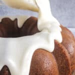 Close-up of a Bundt cake being drizzled with thick white icing, set on a beige plate against a gray background.