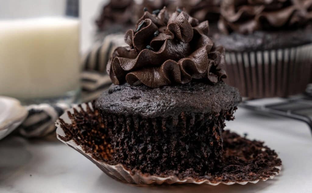 Close-up of a chocolate cupcake with chocolate frosting, partly unwrapped from its liner, and a bite taken out of it. A glass of milk and additional cupcakes are visible in the background.