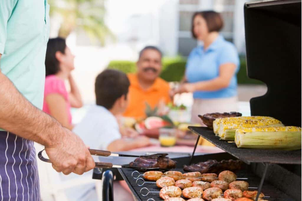 A person grills shrimp, meat, and corn as a family sits and chats at a table in the background, enjoying the backyard BBQ menu.