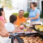 A person grills shrimp, meat, and corn as a family sits and chats at a table in the background, enjoying the backyard BBQ menu.