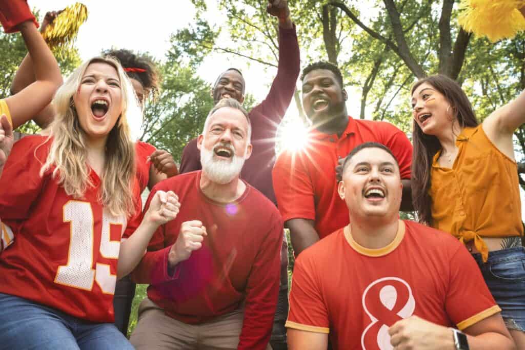 A group of people wearing red sports apparel cheer outdoors with trees in the background.