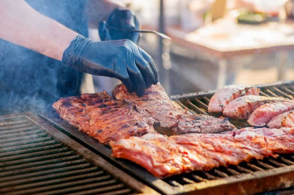 A person wearing black gloves is expertly grilling various cuts of meat, including ribs and steak, on an outdoor barbecue. For those seeking grilling tips for beginners, this setup showcases the importance of proper gear and attention to detail in mastering the art of barbecue.