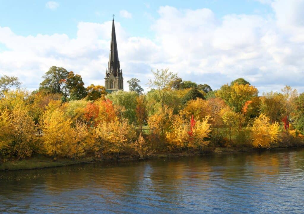 A church with a tall steeple stands behind a line of trees with vibrant autumn foliage by a calm river under a partly cloudy sky.