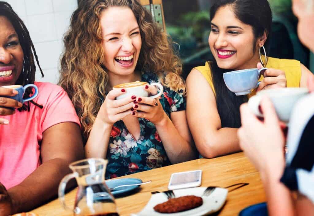 Four people are sitting at a wooden table, laughing and enjoying coffee together. There is a plate with a pastry and a pitcher of coffee on the table.